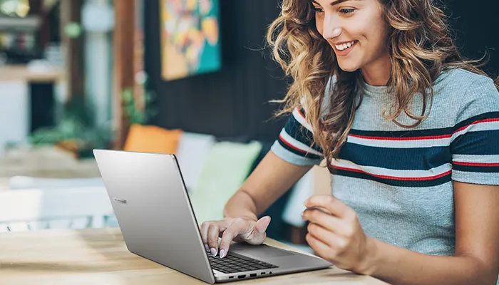 Donna sorride mentre usa un laptop grigio su un tavolo. Indossa una maglietta a righe blu, rosse e grigie. Sullo sfondo si vedono cuscini.