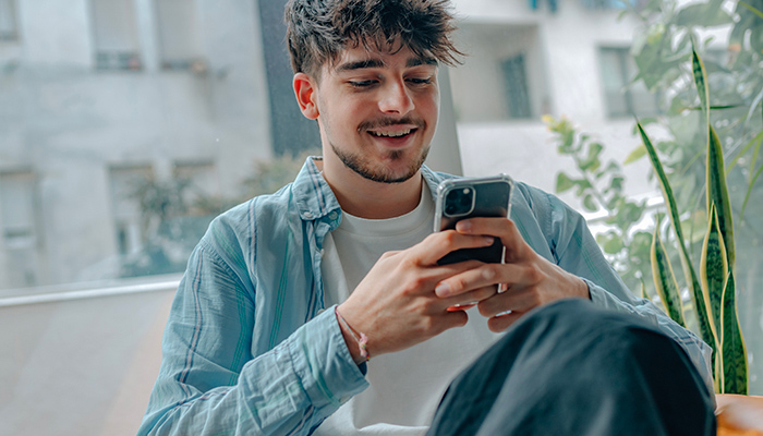 Uomo seduto con camicia azzurra usa un telefono. Dietro, piante e finestre luminose creano uno sfondo rilassante.