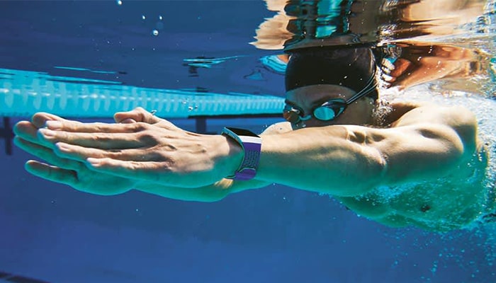 Nuotatore in piscina blu con cuffia, occhialini e orologio al polso. Le mani sono distese in avanti.