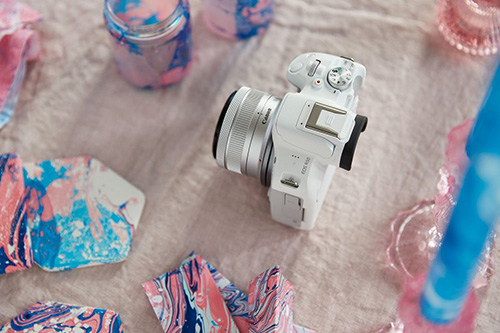 Fotocamera bianca su tessuto con oggetti decorativi rosa e blu. Vasi e ciotole sono sparsi attorno.