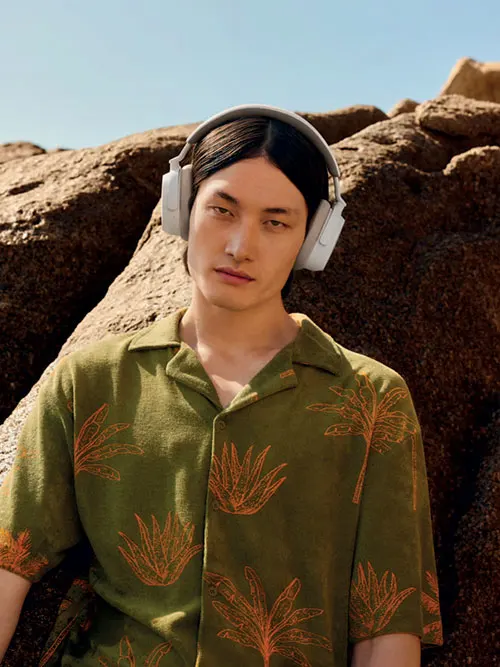Un uomo con cuffie bianche indossa una camicia verde con stampe di palme arancioni, su sfondo di rocce e cielo azzurro.