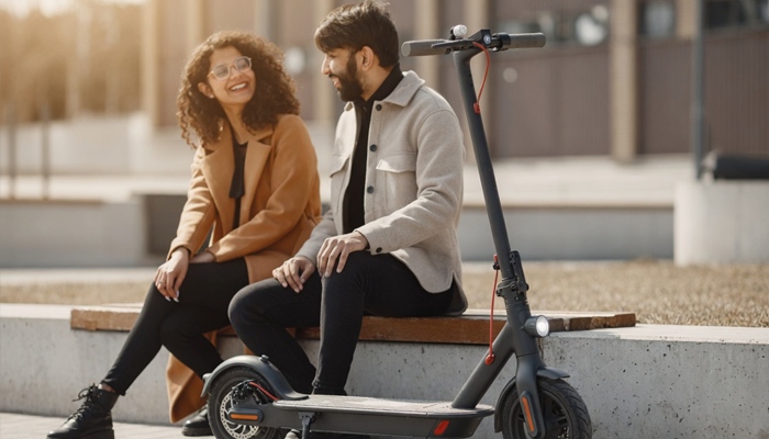 Una donna e un uomo chiacchierano e sorridono seduti su una panchina all'aperto, con un monopattino elettrico nero parcheggiato in primo piano.