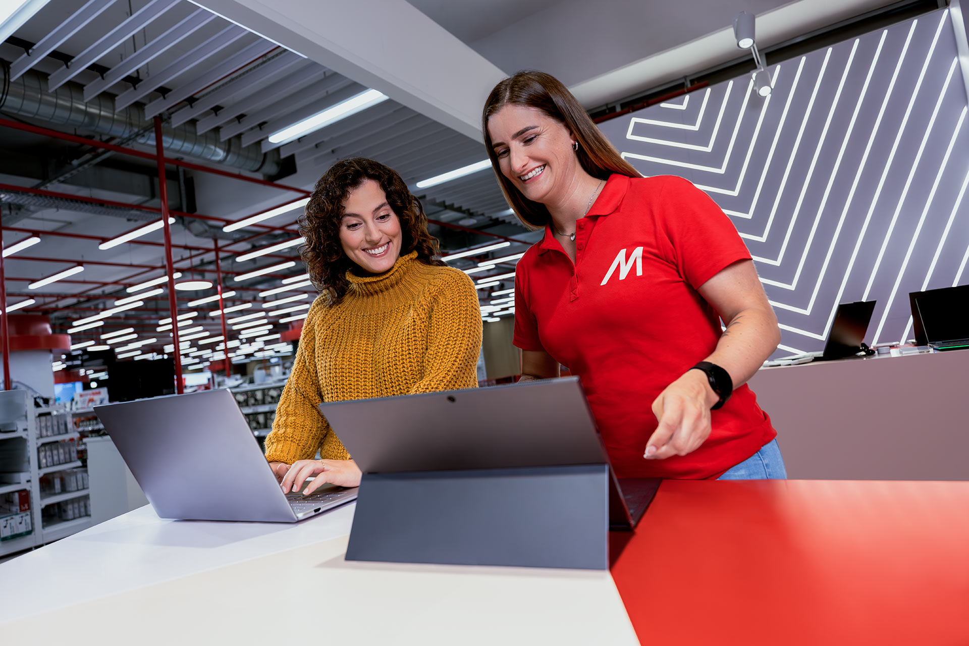 Due donne sorridono davanti a due computer portatili in un negozio di elettronica. La commessa indossa una polo rossa.