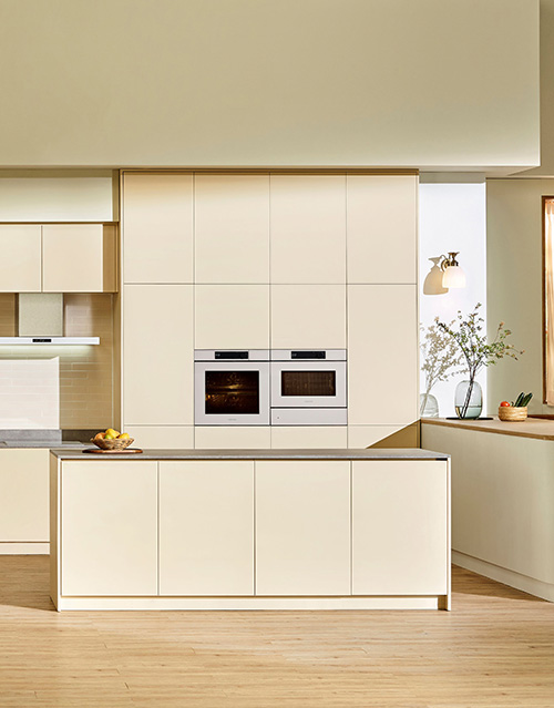 Cucina beige con isola centrale, armadi a muro e forni incassati. Vaso con fiori su ripiano vicino alla finestra. Pavimento in legno.