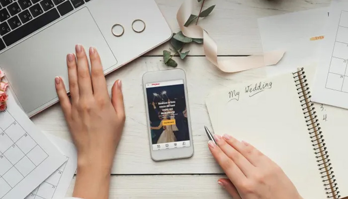 Vista dall'alto di una scrivania con laptop, anelli, telefono, agenda con la scritta "My Wedding", calendario e mani.