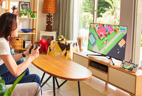 Donna seduta gioca con controller. Tavolo di legno, TV con videogioco, mobile TV e finestra.