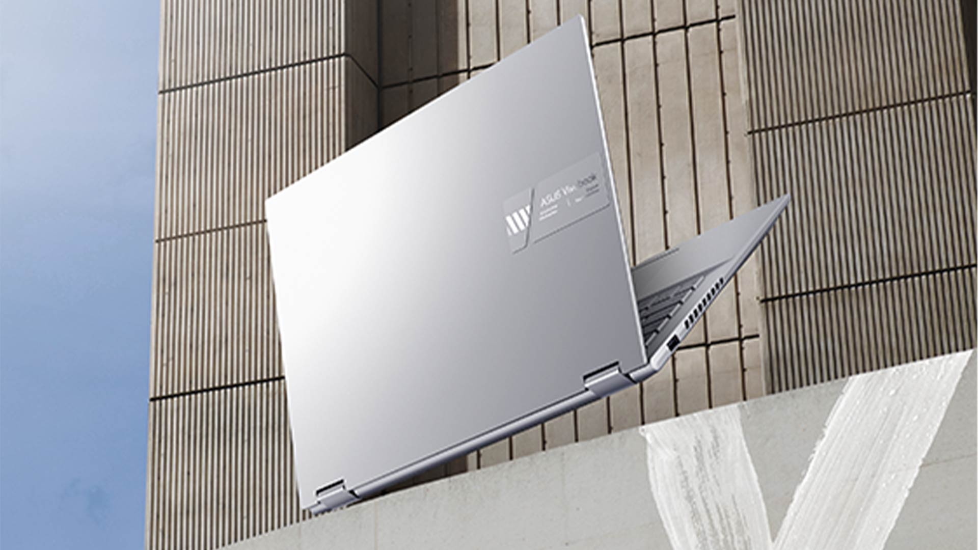 Un computer portatile grigio aperto è appoggiato su un muro di pietra. Il muro ha delle strisce verticali. Il cielo è visibile.