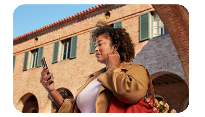 Donna con auricolari e smartphone, indossa giacca marrone e maglietta bianca. Dietro, edificio beige con finestre verdi e cielo azzurro.