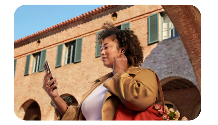 Donna con auricolari e smartphone, indossa giacca marrone e maglietta bianca. Dietro, edificio beige con finestre verdi e cielo azzurro.