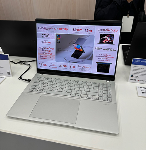 Un laptop aperto su un tavolo mostra specifiche tecniche sullo schermo. Tastiera e touchpad grigi visibili.