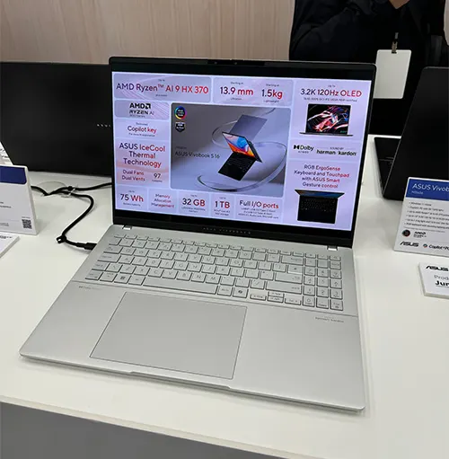 Un laptop aperto su un tavolo mostra specifiche tecniche sullo schermo. Tastiera e touchpad grigi visibili.
