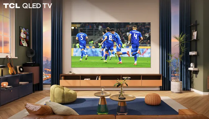 Soggiorno con TV che mostra una partita di calcio, divano in pelle marrone, tavolini e piante. Tende blu ai lati della TV.