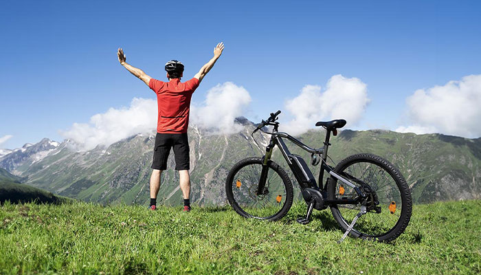Ciclista in maglia rossa alza le braccia vicino a bici nera su prato verde, con montagne sullo sfondo.