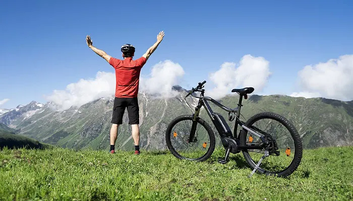 Ciclista in maglia rossa alza le braccia vicino a bici nera su prato verde, con montagne sullo sfondo.