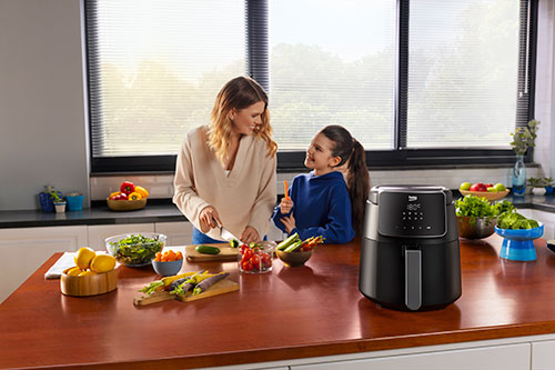 Una donna taglia verdure su un piano di lavoro marrone in cucina, vicino a una friggitrice ad aria nera e a una ragazza.