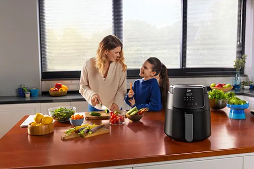 Una donna taglia verdure su un piano di lavoro marrone in cucina, vicino a una friggitrice ad aria nera e a una ragazza.