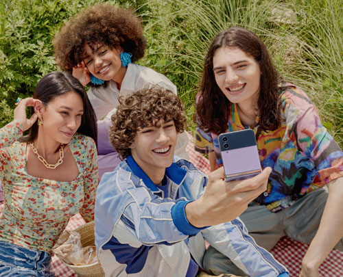Quattro persone sorridono in un picnic. Un ragazzo tiene un telefono viola e scatta una foto. Cesto da picnic e vegetazione sullo sfondo.