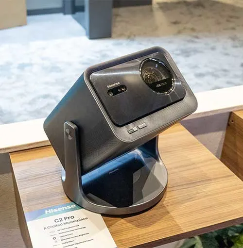 Proiettore grigio su base curva, appoggiato su un tavolo di legno chiaro. Brochure bianca visibile in primo piano.