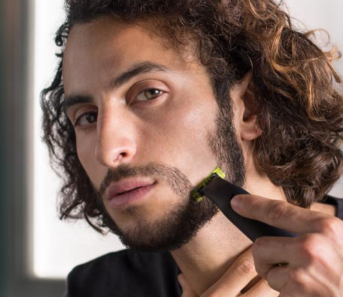 Un uomo con i capelli ricci usa un rasoio nero e verde per modellare la barba. Sullo sfondo c'� una finestra.
