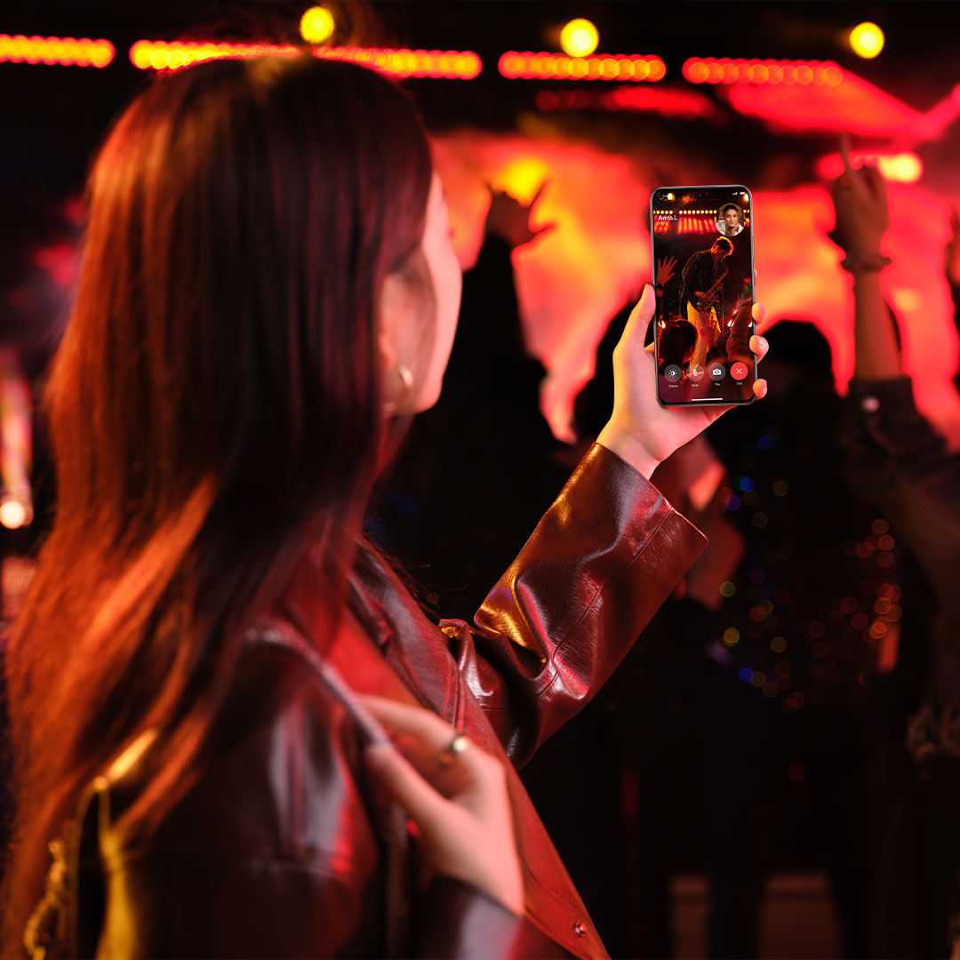 Donna in giacca tiene un telefono con una videochiamata in un locale affollato. Luci rosse e gialle sullo sfondo.