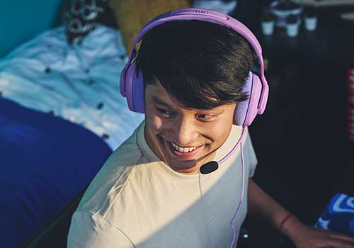 Uomo sorridente con cuffie viola e microfono. Indossa una t-shirt bianca. Sullo sfondo, un letto con cuscini e oggetti su una scrivania.