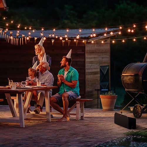 Famiglia con cappelli da festa seduta a tavola sotto luci esterne. Grill e altoparlante visibili sullo sfondo.