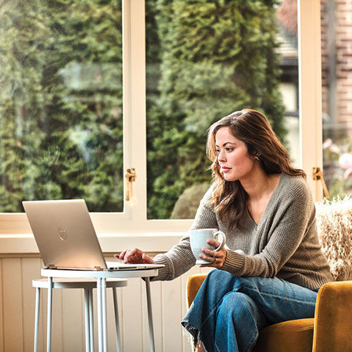 Donna seduta su una sedia gialla con laptop su tavolino argento, tiene una tazza bianca. Grande finestra con vista su alberi.