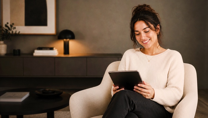 Una donna sorridente è seduta su una poltrona mentre guarda uno schermo di un tablet che tiene tra le mani, in un soggiorno dal design moderno.