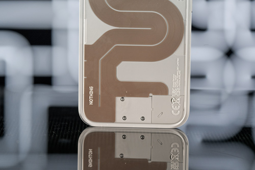 Dettaglio del retro di un telefono cellulare con design geometrico marrone e scritta "NOTHING" su sfondo sfocato.