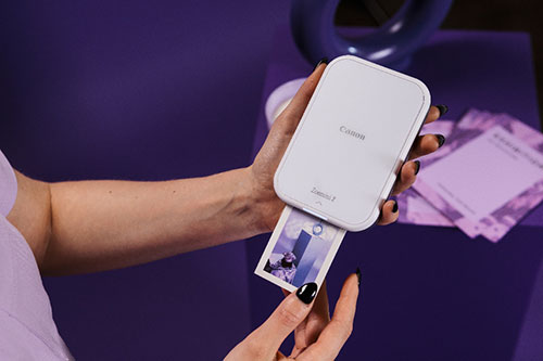 Mani tengono una stampante portatile bianca che stampa una foto su sfondo viola, con confezioni di carta fotografica visibili.