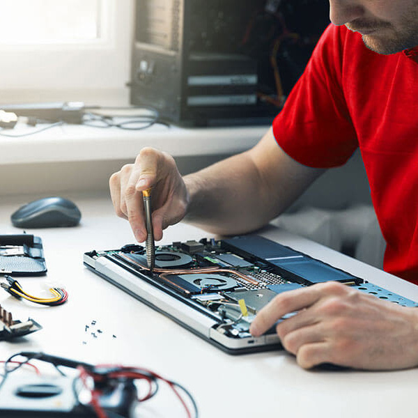Un tecnico ripara un laptop aperto con un cacciavite su un tavolo bianco, con componenti sparsi e un computer sullo sfondo.