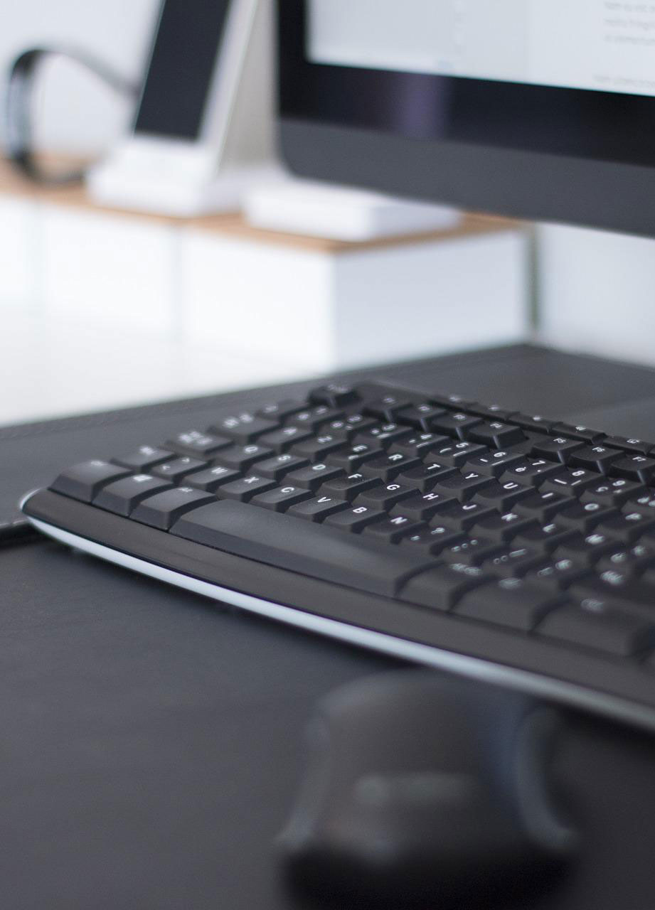 Scrivania nera con tastiera, mouse e monitor del computer. Sullo sfondo, telefono cellulare e cuffie su una superficie bianca.