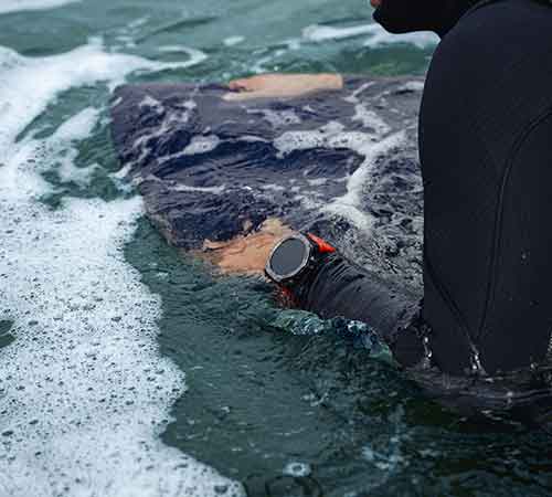 Una persona in mare indossa una muta nera e un orologio da polso con cinturino rosso. L'acqua è agitata con schiuma bianca.