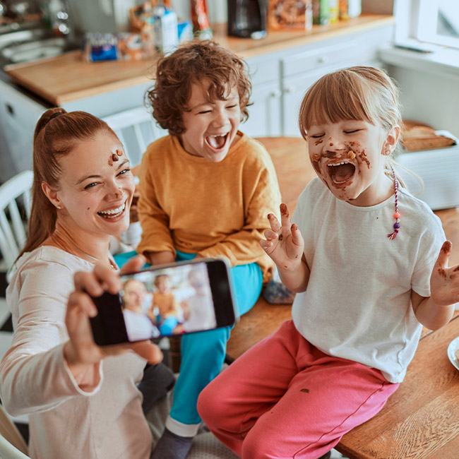 Donna sorridente fa selfie con bambini sporchi di cioccolato su tavolo in cucina. Uno indossa maglia gialla, l'altro rosa.