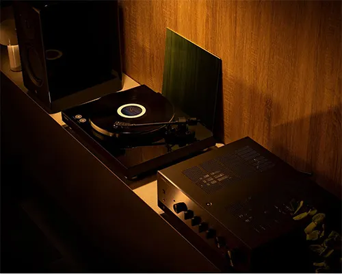 Un giradischi nero con disco in vinile su un mobile di legno, accanto a un amplificatore, su sfondo di legno.