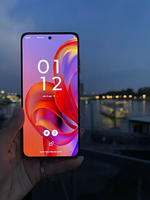 Una mano tiene un telefono che mostra l'ora 01:12. Sullo sfondo si vede un fiume e delle luci.