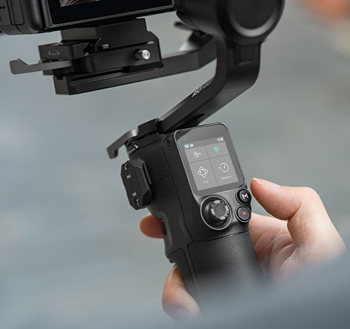 Una mano tiene in mano un gimbal nero con schermo acceso e pulsanti di controllo, su sfondo grigio.
