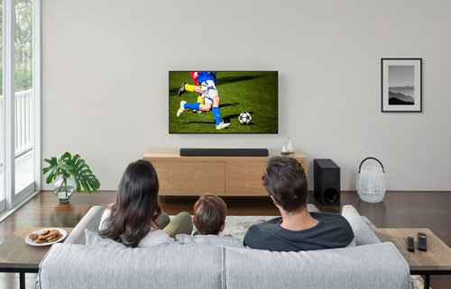 Famiglia seduta sul divano guarda una partita di calcio in TV. Tavolino con telecomandi e piatto di biscotti.