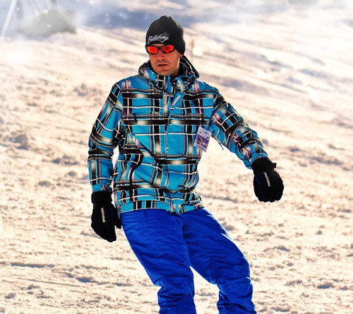 Un uomo con cappello e occhiali da sole indossa una giacca a quadri blu e pantaloni blu su una collina innevata.