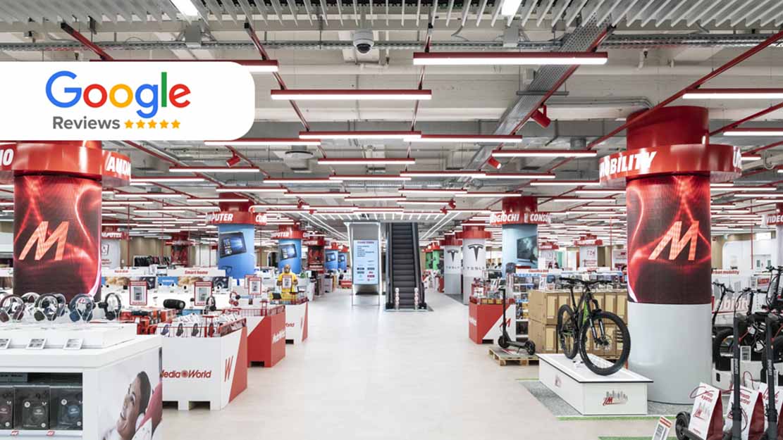 Un grande negozio con scaffali bianchi e rossi pieni di elettronica. Al centro, scale mobili e insegna "Google Reviews".