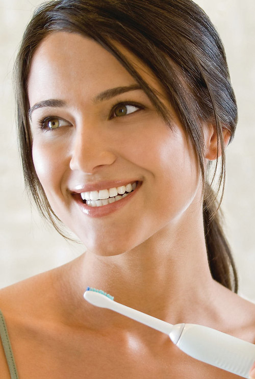 Donna sorridente tiene uno spazzolino bianco con setole blu, sullo sfondo un muro chiaro.