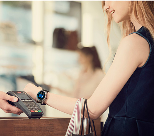 Donna paga con smartwatch avvicinando il polso a un terminale di pagamento grigio, con borsa a righe. Vetrine sullo sfondo.