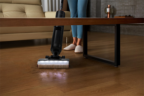 Una persona pulisce un pavimento di legno con un aspirapolvere. Un divano e un tavolino sono sullo sfondo.