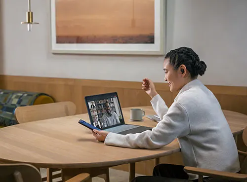 Donna seduta a un tavolo di legno, con laptop che mostra una videochiamata e uno smartphone in mano. Tazza sul tavolo.