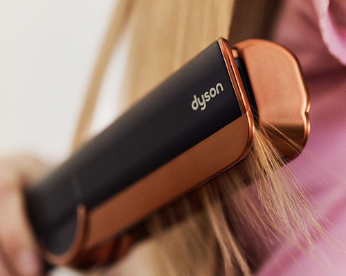 Piastra Dyson nera e rame che modella ciocche. Il marchio Dyson � visibile sul corpo nero. Sfondo sfocato rosa e beige.