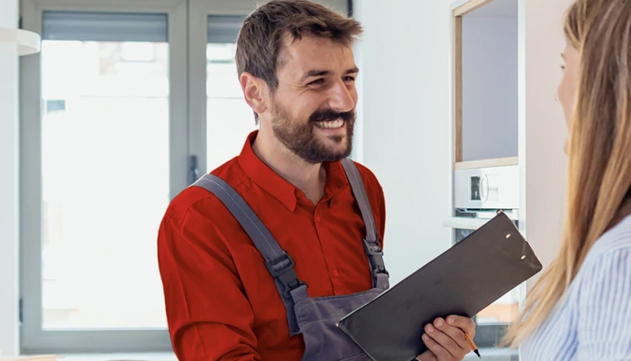 Uomo sorridente in tuta rossa e grigia con blocco appunti nero parla con donna in camicia a righe in interno luminoso.