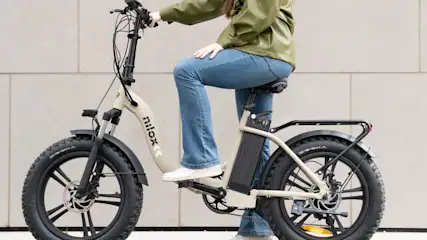 Donna su bici elettrica beige con pneumatici neri larghi. Indossa jeans blu e giacca verde. Sfondo di muro grigio.