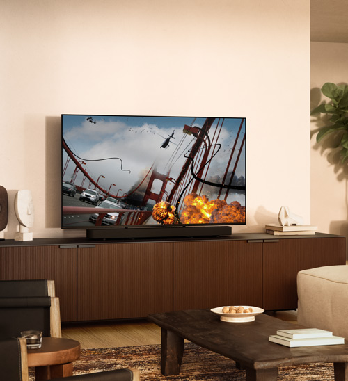 Soggiorno con TV che mostra un ponte in fiamme, auto, elicottero e esplosioni su mobile marrone. Tavolino da caffè di fronte.