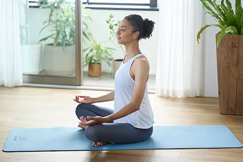Donna in posizione yoga su tappetino blu. Indossa canotta bianca e pantaloni grigi. Piante e finestre sullo sfondo.