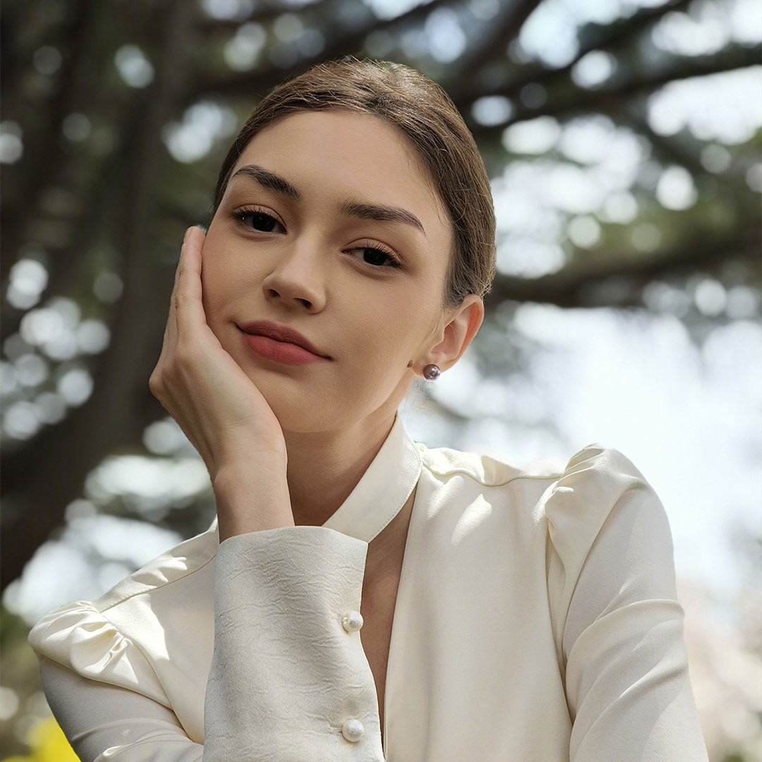 Donna con camicia bianca, mano sul viso, sfondo alberi. Bottoni di perla sulla manica.